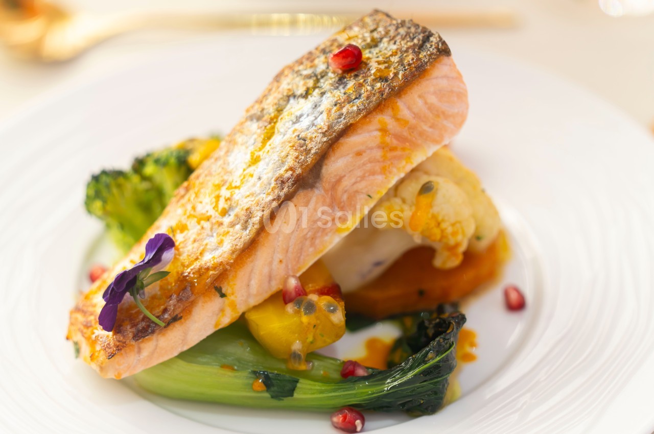Filet de saumon grillé servi avec légumes variés, purée et graines de grenade sur une assiette blanche.