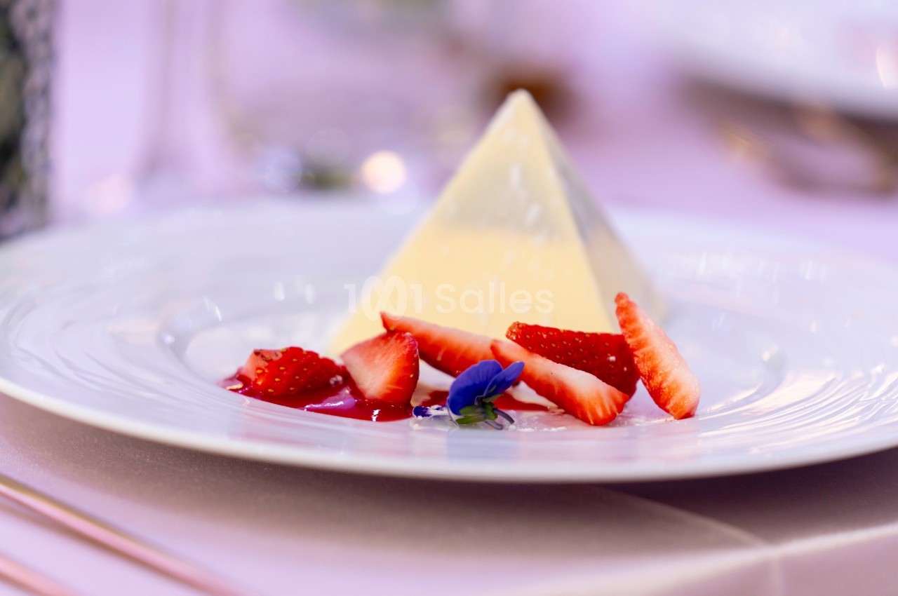 Dessert pyramidal en fromage blanc accompagné de fraises tranchées et d'une fleur comestible sur une assiette blanche.