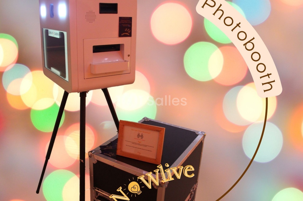 Borne photobooth blanche sur trépied devant un fond flou coloré, accompagnée d'une valise noire.