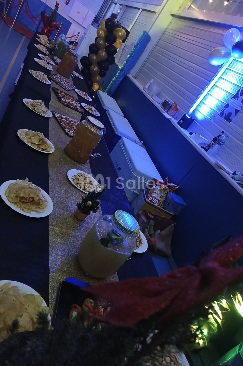 Table décorée avec nappes noires et dorées, garnie de plats variés et boissons, dans une salle éclairée par des lumières…