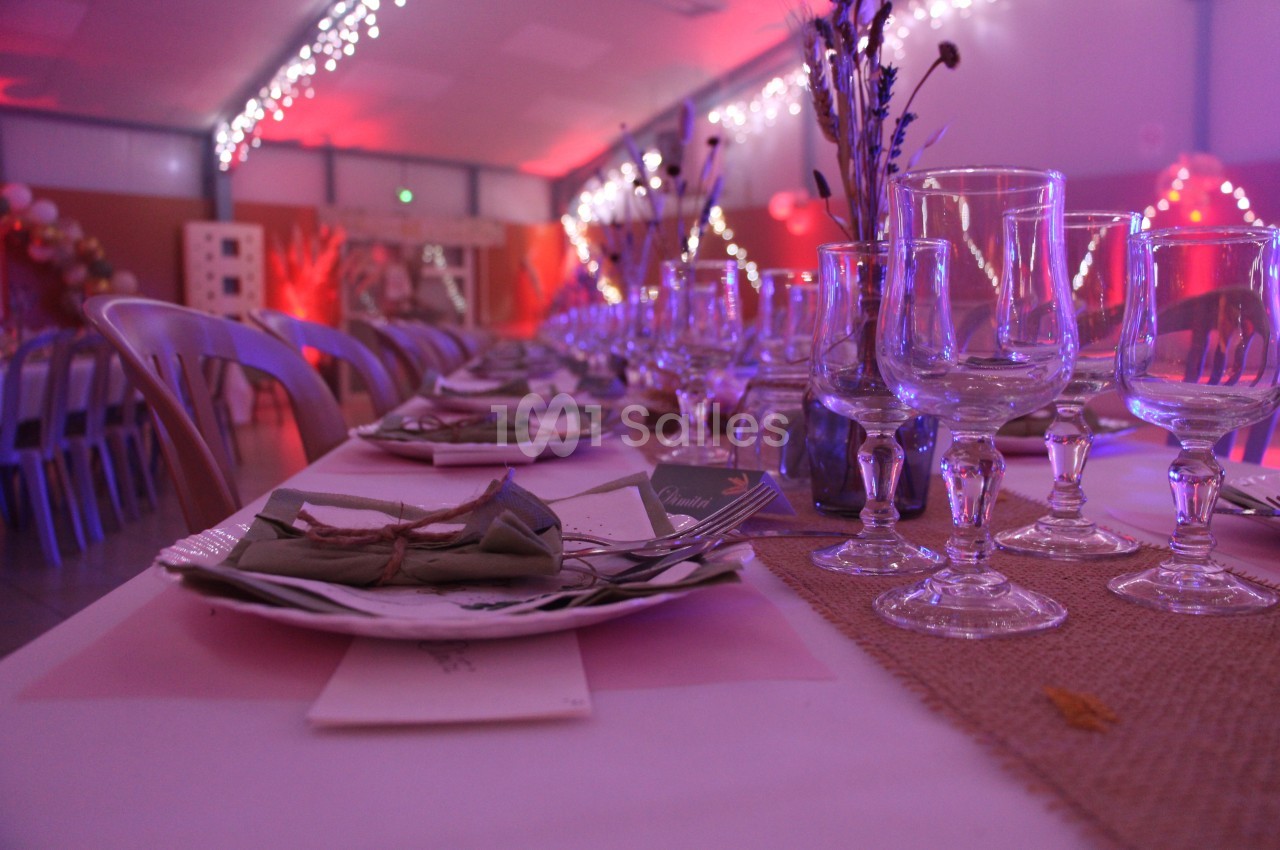 Table décorée pour un événement, avec nappes blanches, verres, couverts et éclairage tamisé aux tons roses.
