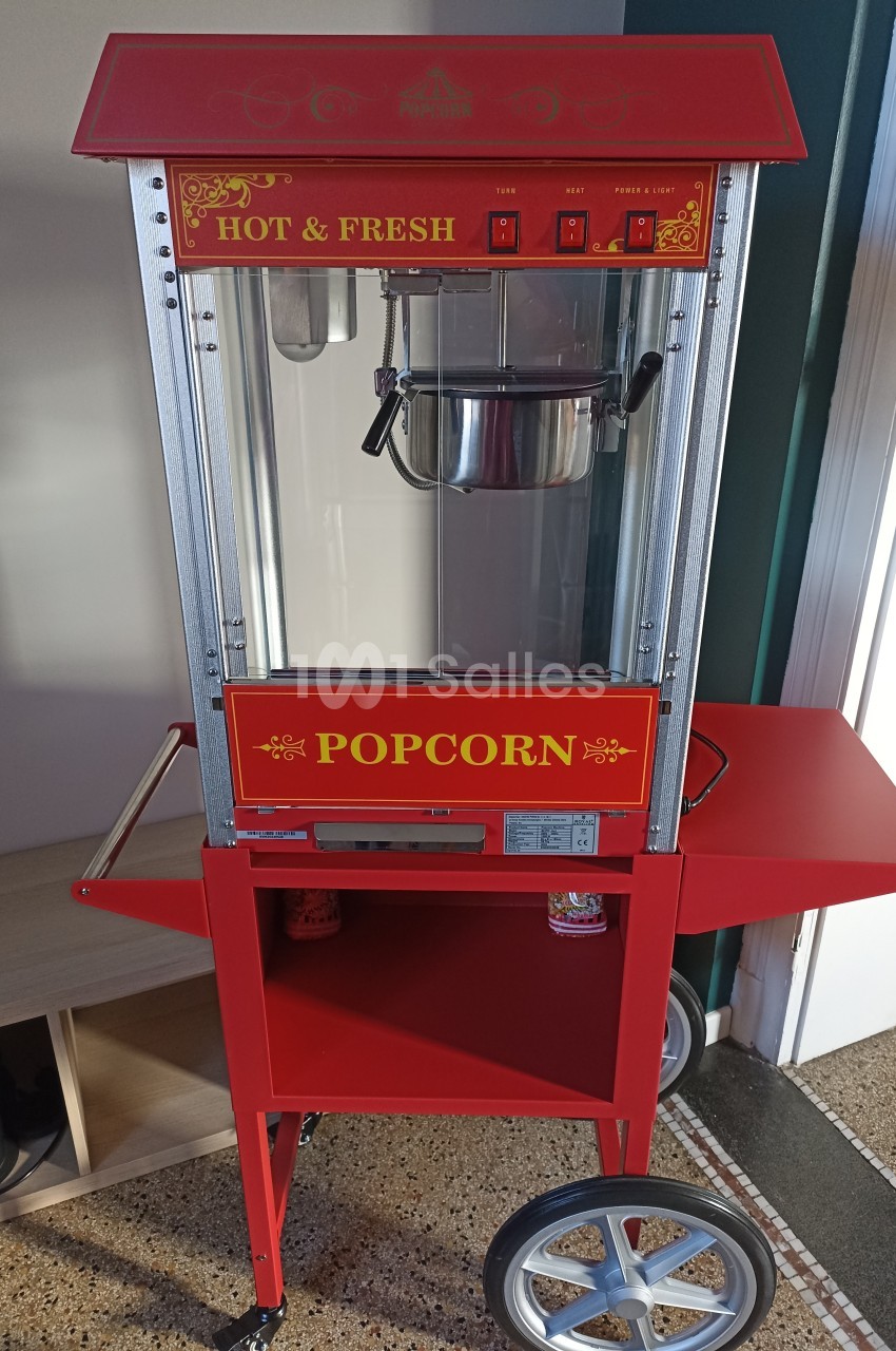 Machine à popcorn rouge sur roulettes, avec compartiment de rangement et inscription ’Hot & Fresh’ sur le haut.