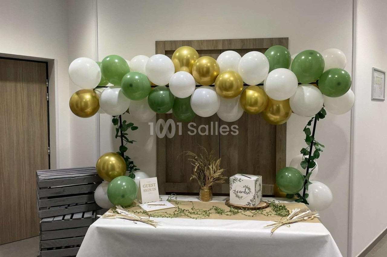 Décoration de table avec ballons verts, blancs et dorés, boîte à cartes et livre d'or dans un espace intérieur.