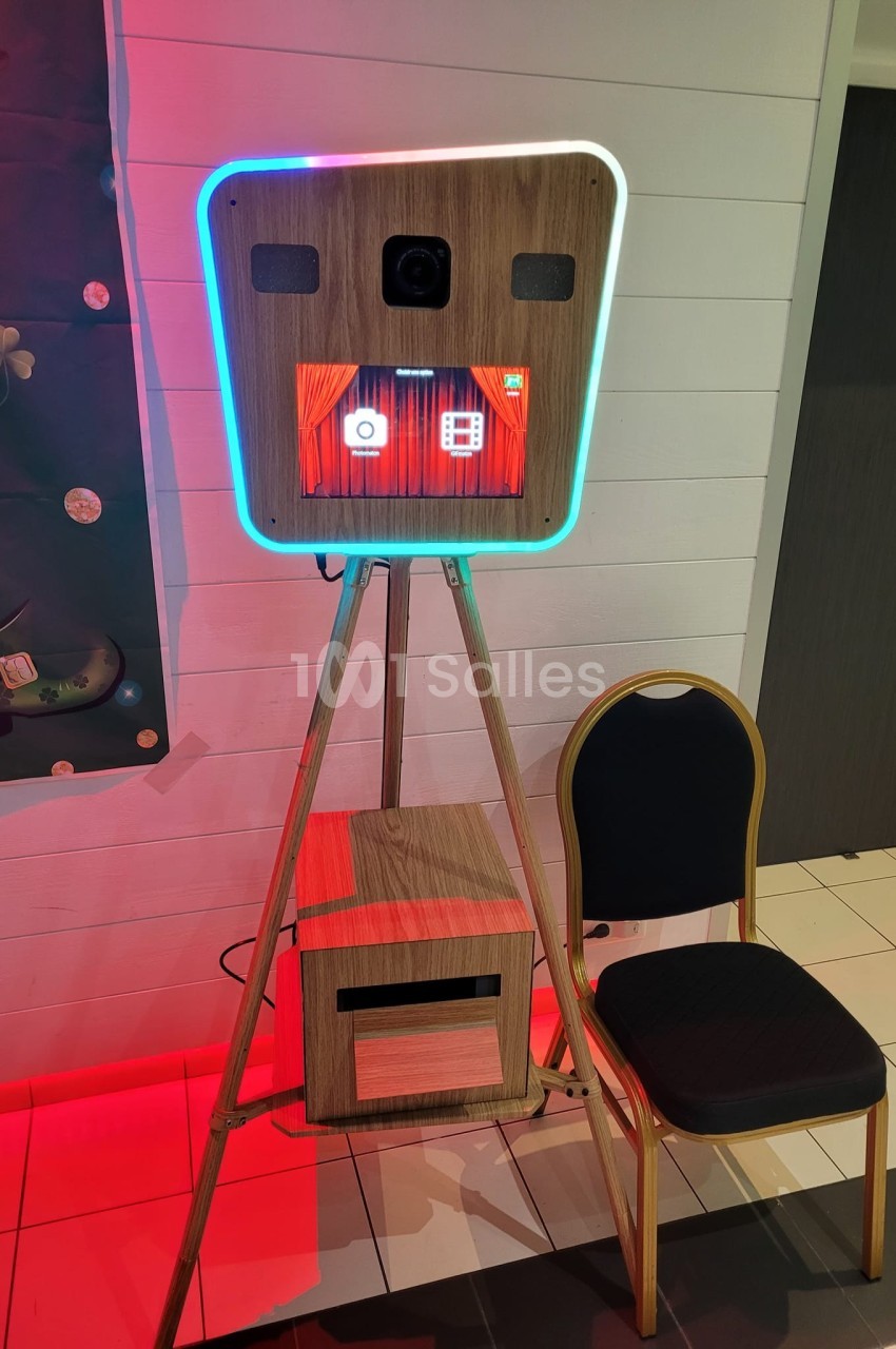 Borne photo interactive en bois avec écran tactile, posée sur un trépied, accompagnée d'une chaise noire.
