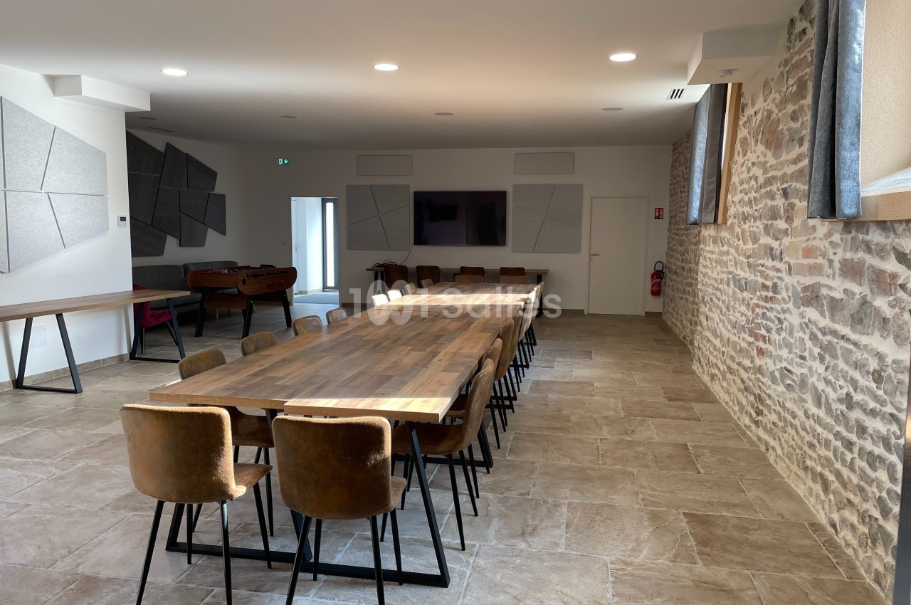 Salle lumineuse avec longues tables en bois, chaises en cuir, murs en pierre et panneaux acoustiques modernes.