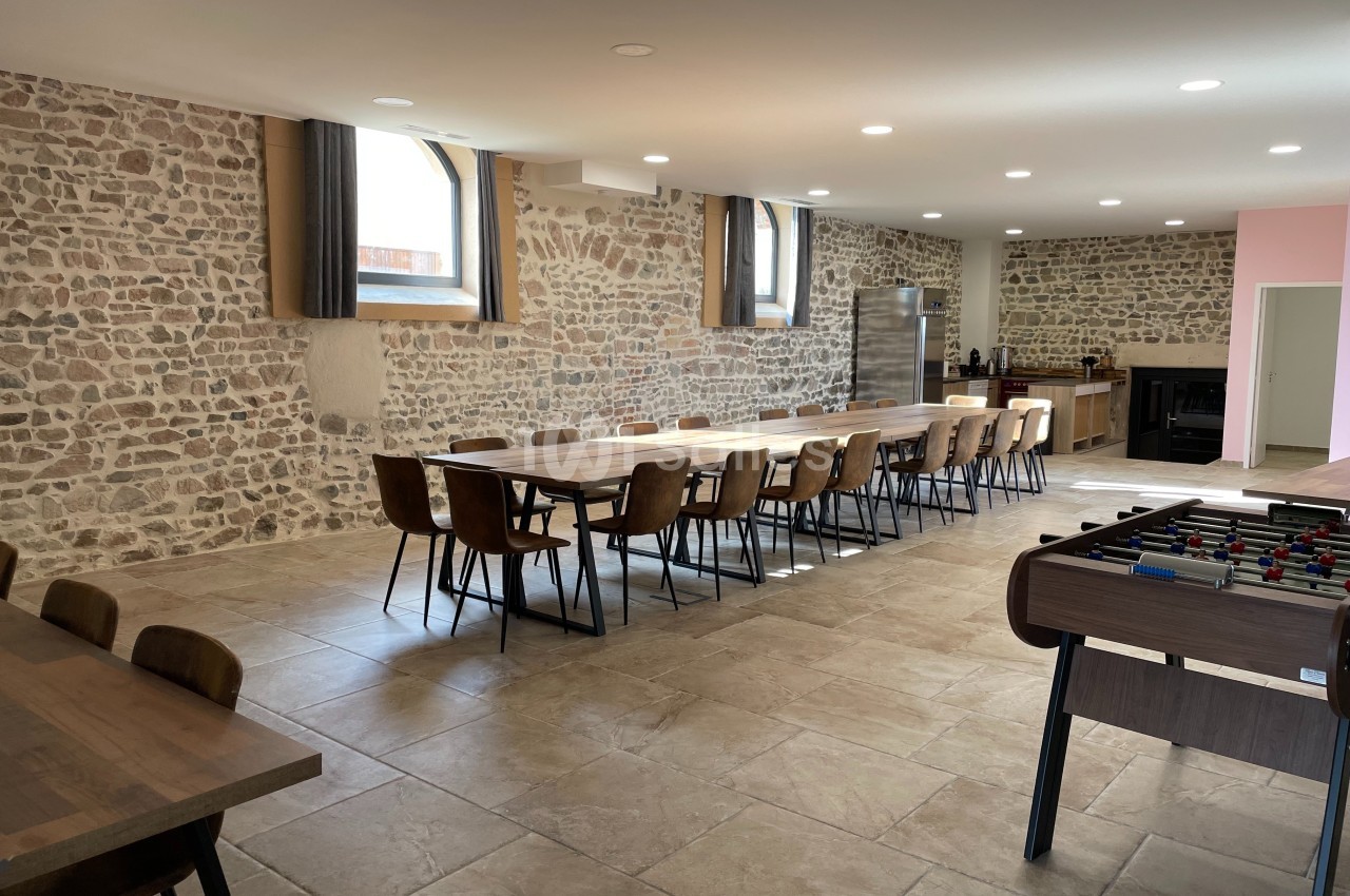 Salle lumineuse avec murs en pierre, grande table en bois entourée de chaises, baby-foot et cuisine en arrière-plan.
