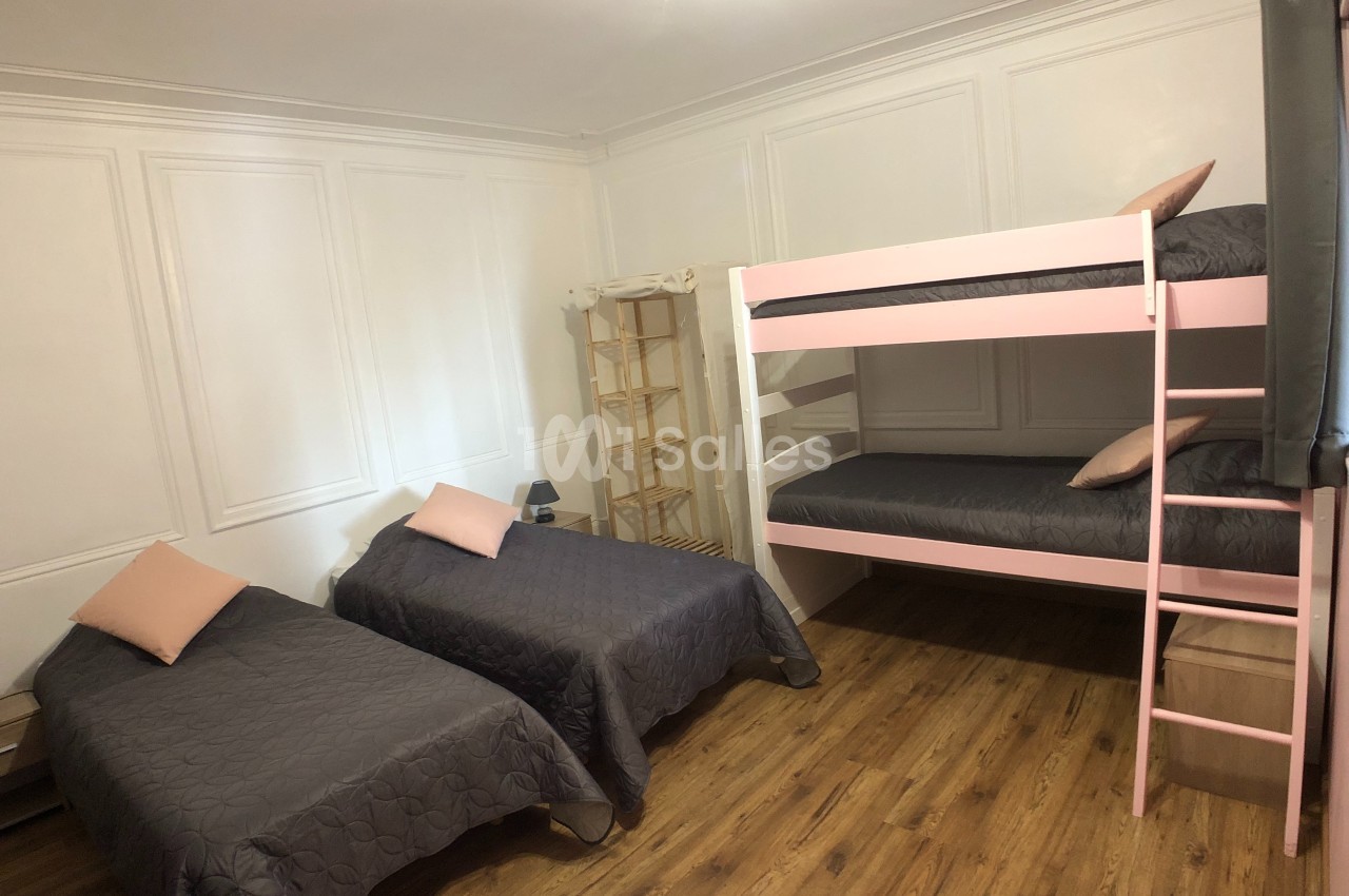 Chambre avec deux lits simples, un lit superposé rose, parquet au sol et murs blancs.