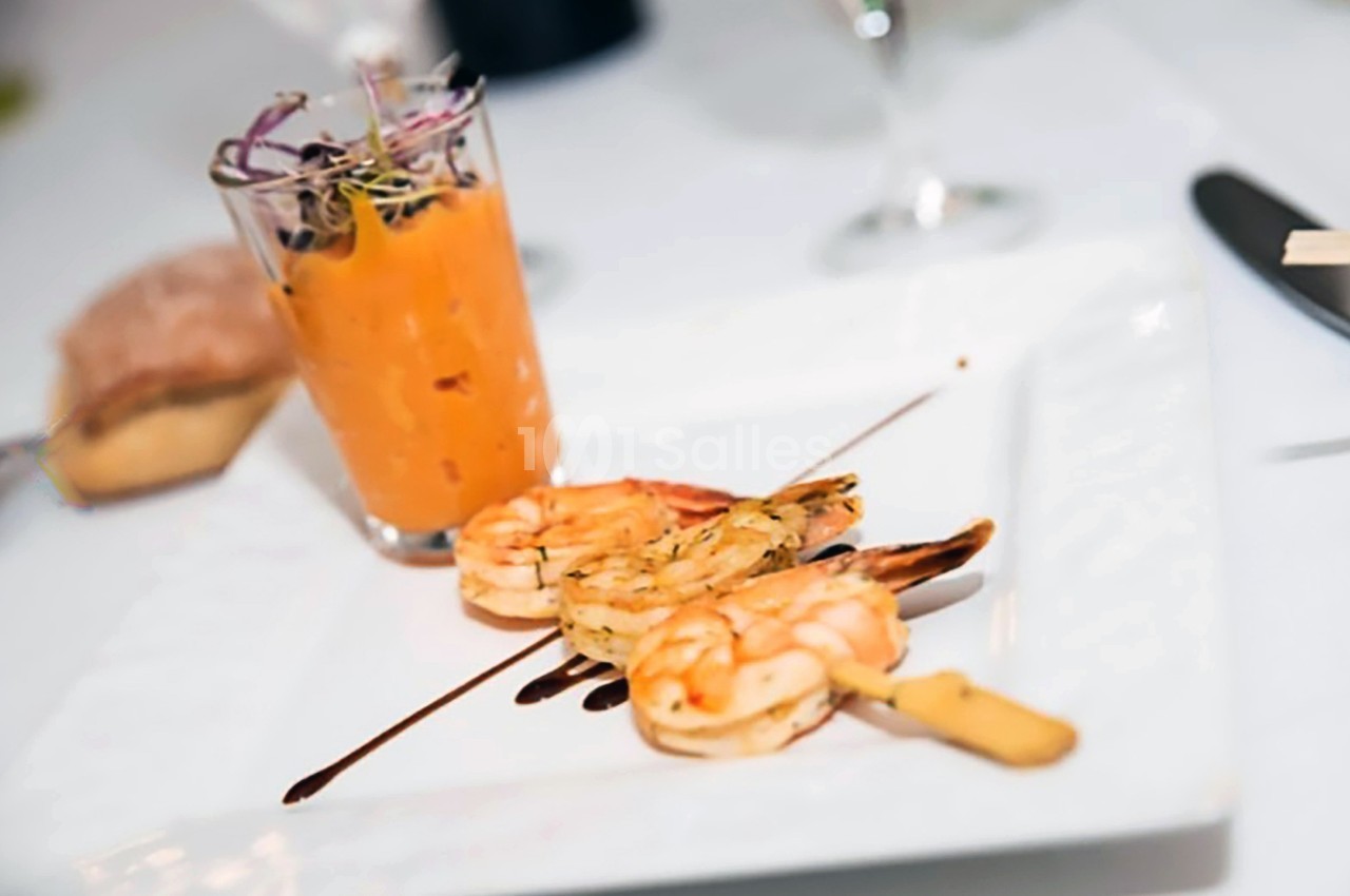 Brochette de crevettes grillées avec sauce balsamique, accompagnée d'un verre de soupe froide et d'une tranche de pain.