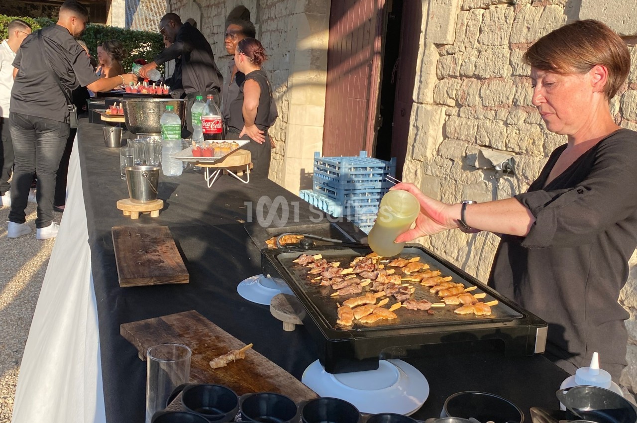 Une femme cuisine des brochettes sur une plancha lors d'un buffet en extérieur, avec des boissons et des plats disposés sur…