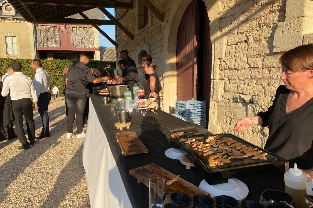 Buffet en extérieur avec des plats chauds préparés sur une plancha, invités discutant près d'un bâtiment en pierre.