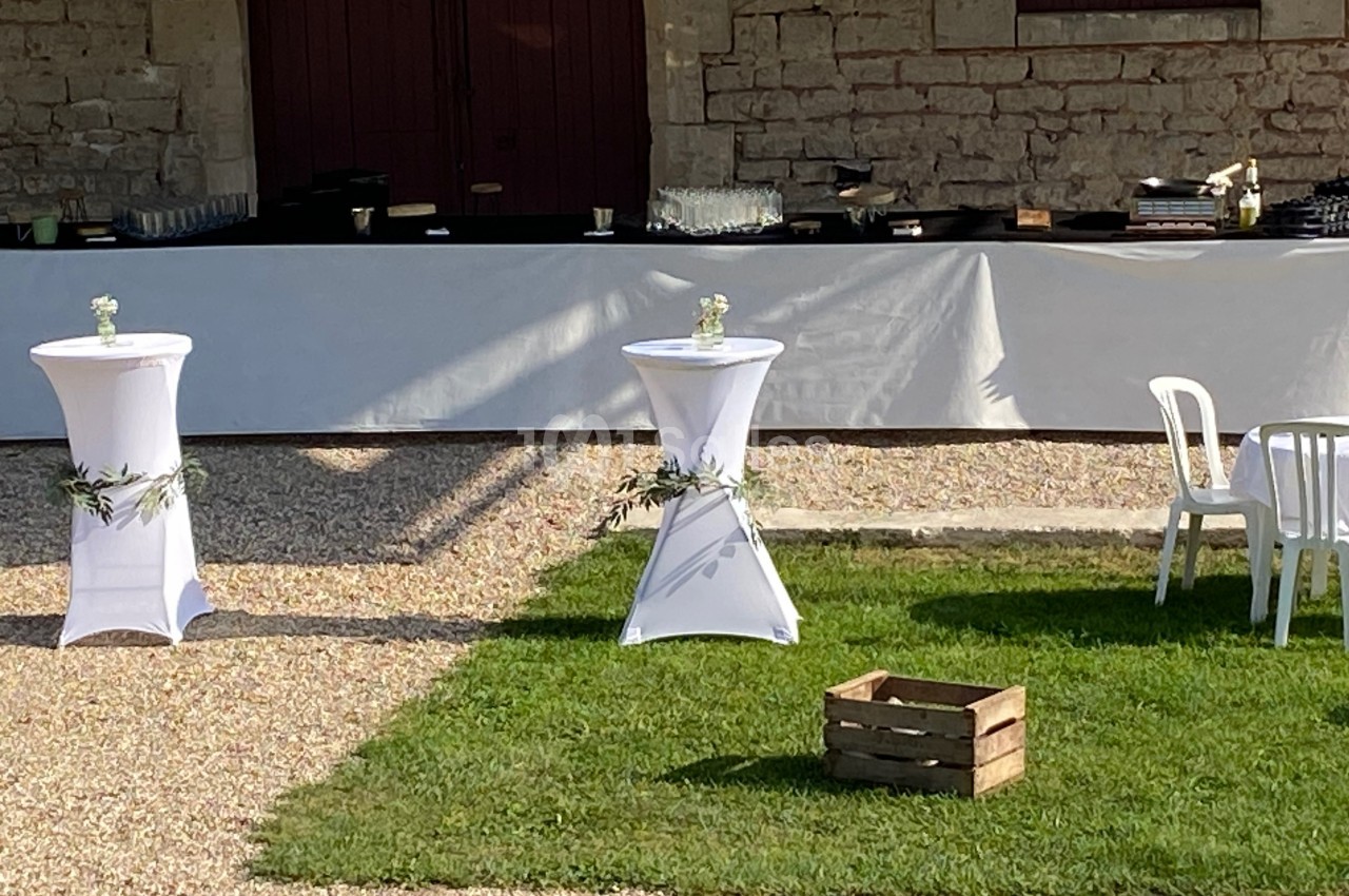 Deux tables hautes décorées de fleurs et de feuillage sur une pelouse, avec un buffet en arrière-plan.