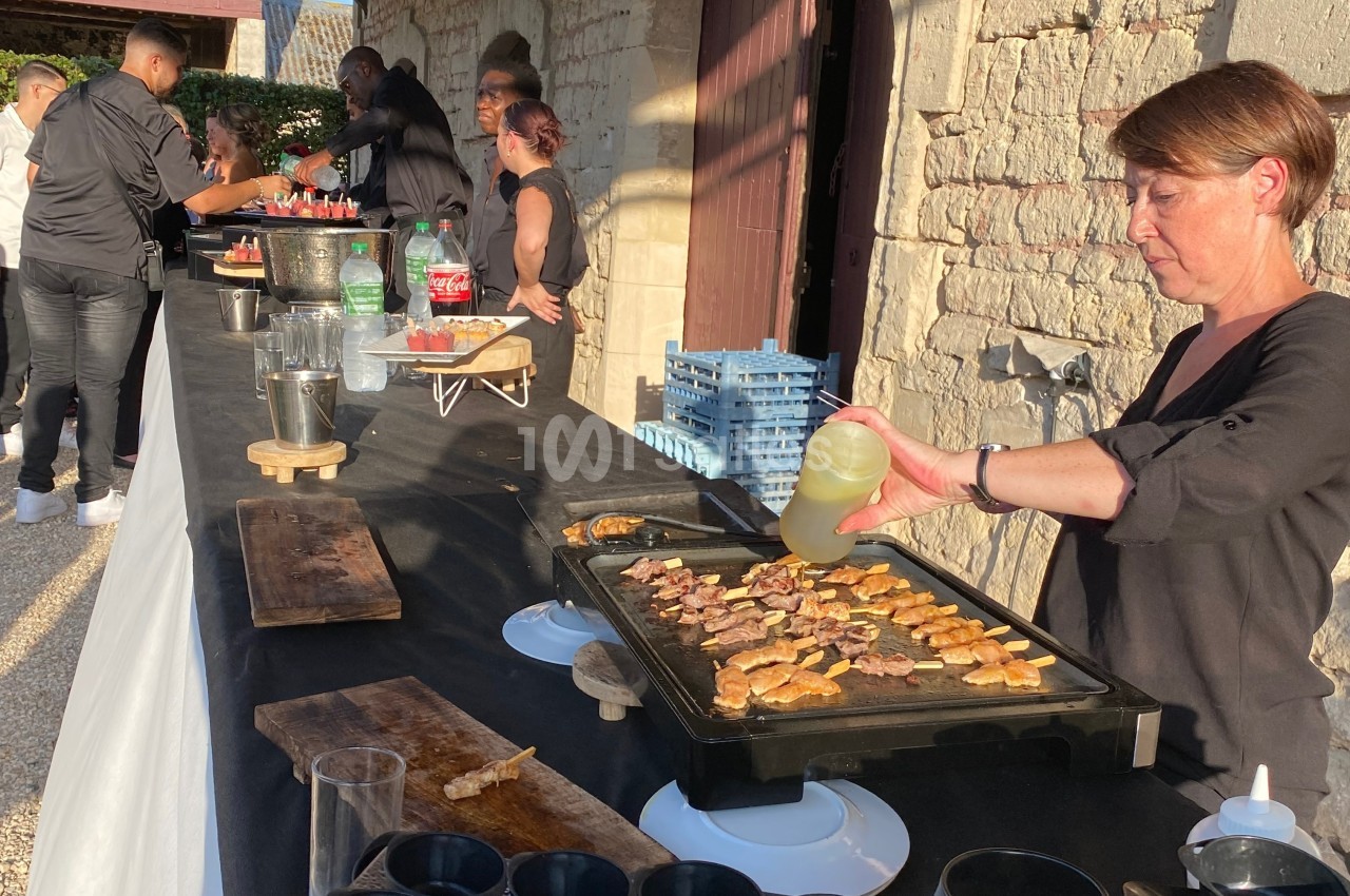 Une femme cuisine des aliments sur une plancha lors d'un buffet en extérieur, avec des serveurs en arrière-plan.