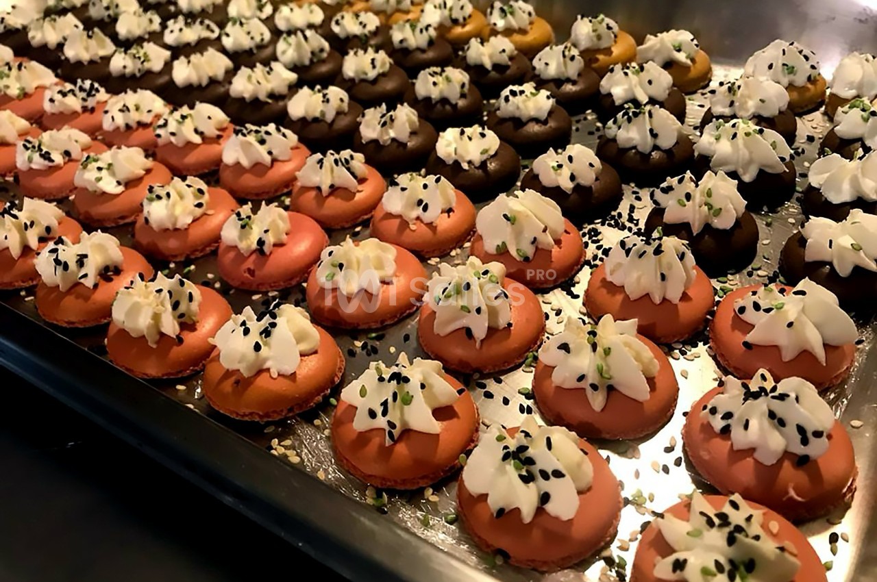 Plateau de petits macarons colorés garnis de crème et parsemés de graines noires et vertes.