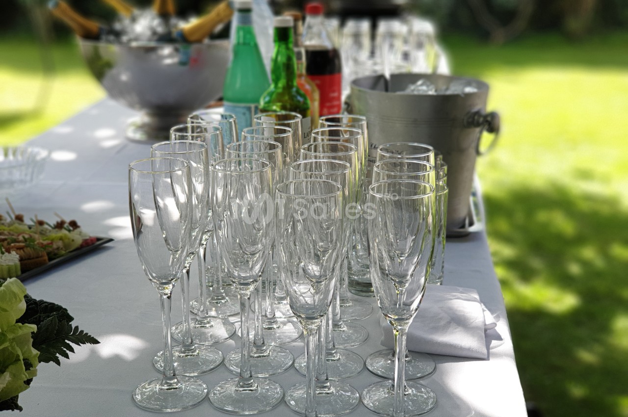 Rangée de flûtes à champagne vides sur une table dressée en extérieur, avec boissons et seau à glace en arrière-plan.