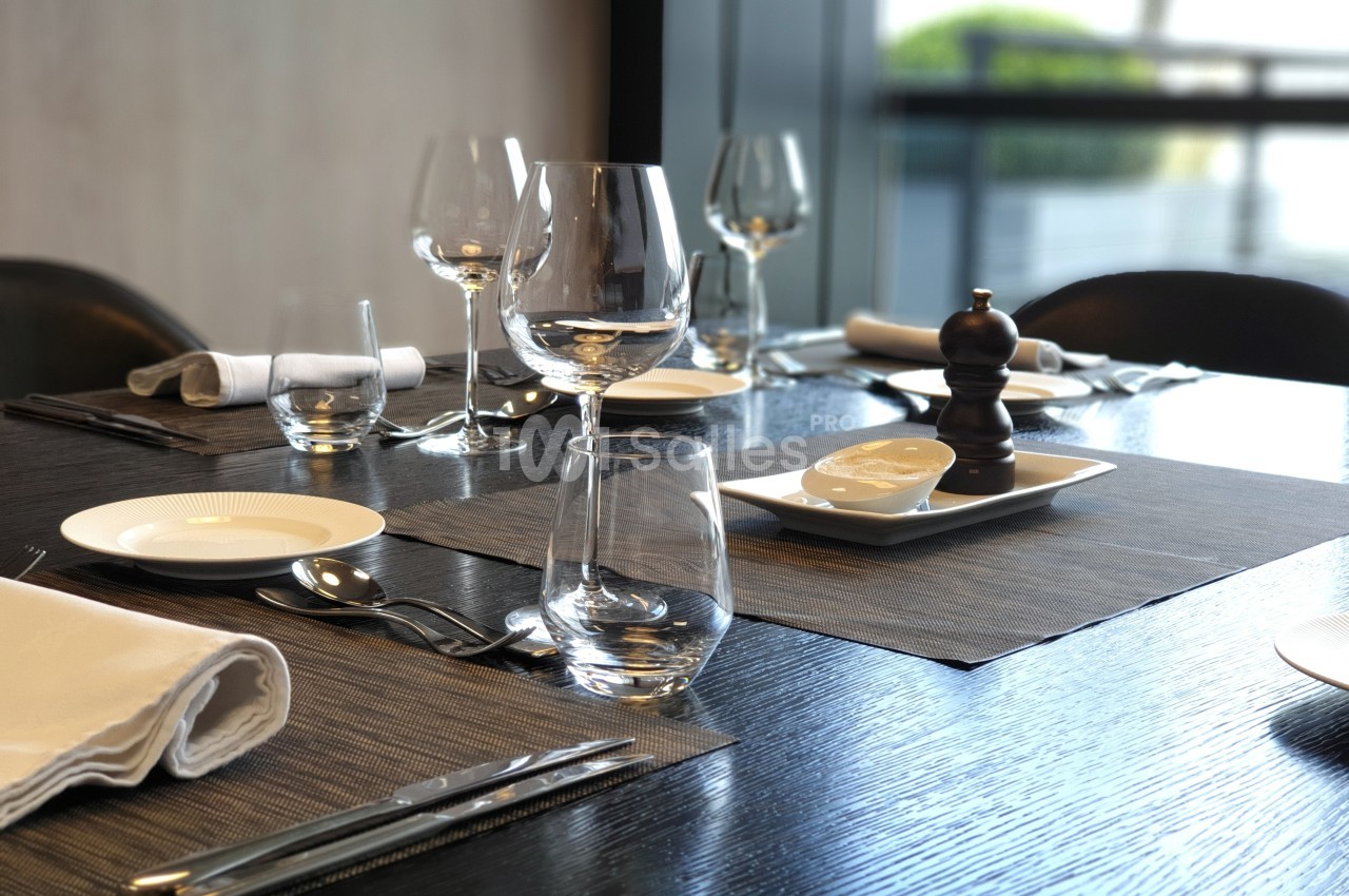 Table dressée avec des verres, assiettes, couverts et serviettes, sur un fond lumineux avec une vue extérieure floue.