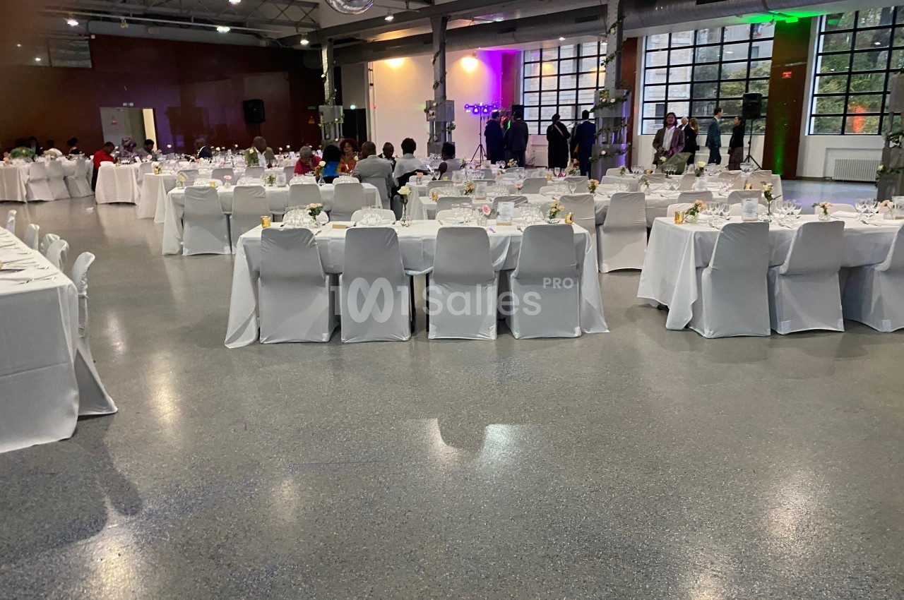 Salle de réception avec tables rondes et rectangulaires dressées, chaises blanches et invités en arrière-plan.