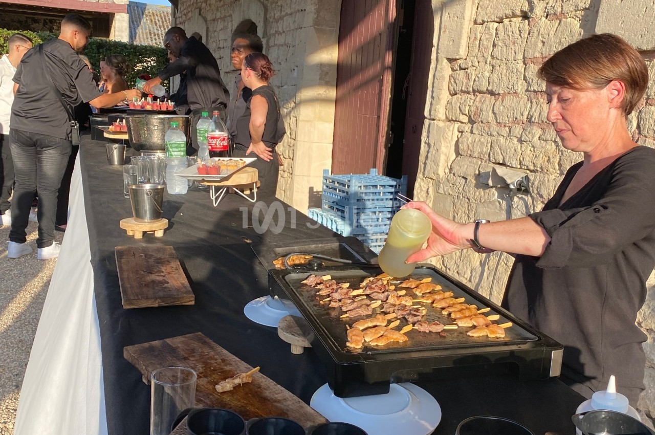 Une femme cuisine des brochettes de viande sur une plancha lors d'un événement en extérieur, avec des serveurs à l'arrière…
