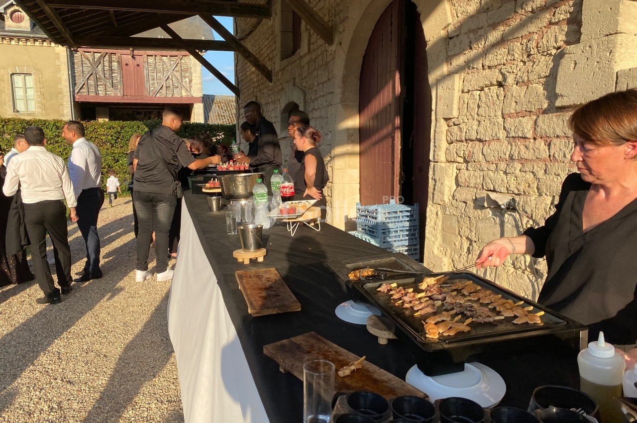 Buffet en extérieur avec des plats chauds préparés sur une plancha, des invités discutent à proximité.