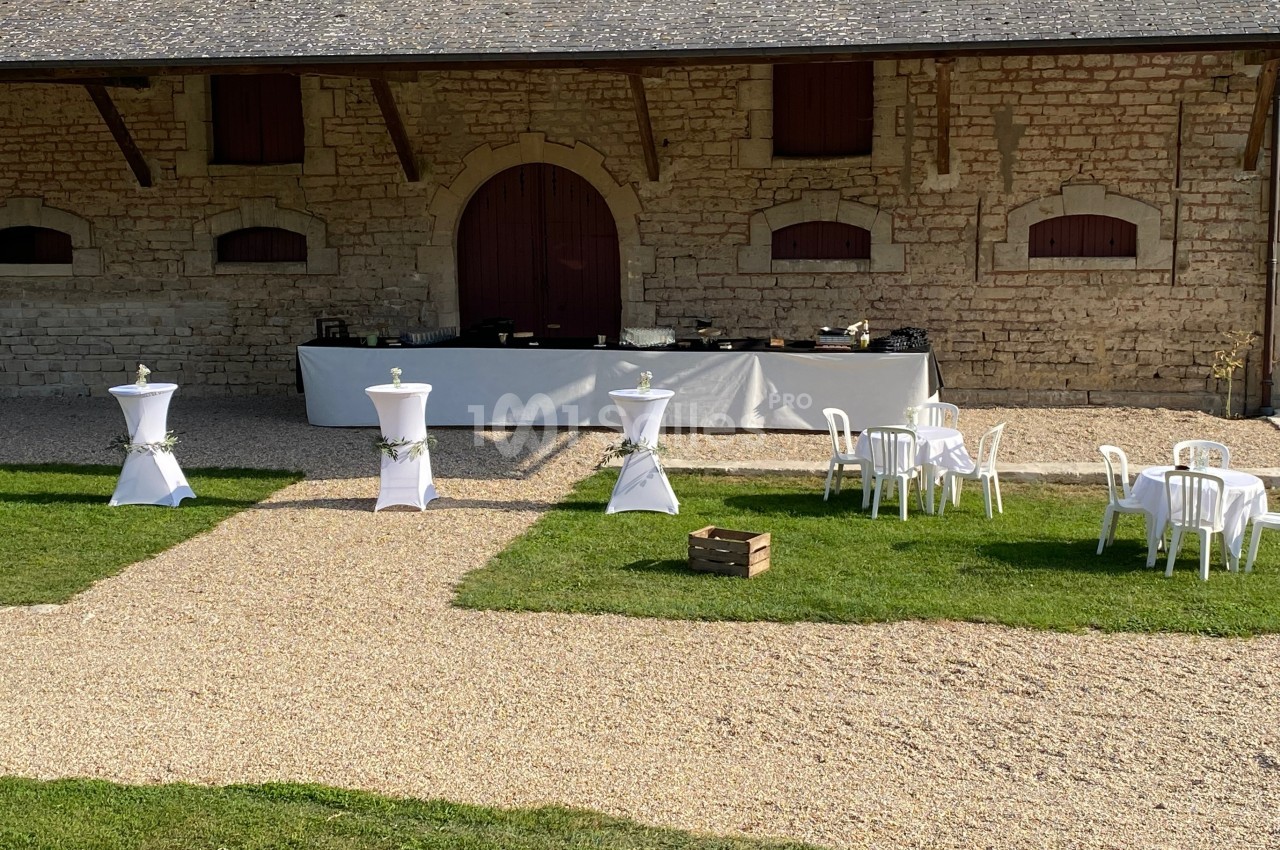 Espace extérieur avec tables et mange-debout dressés devant un bâtiment en pierre, décoré pour un événement.