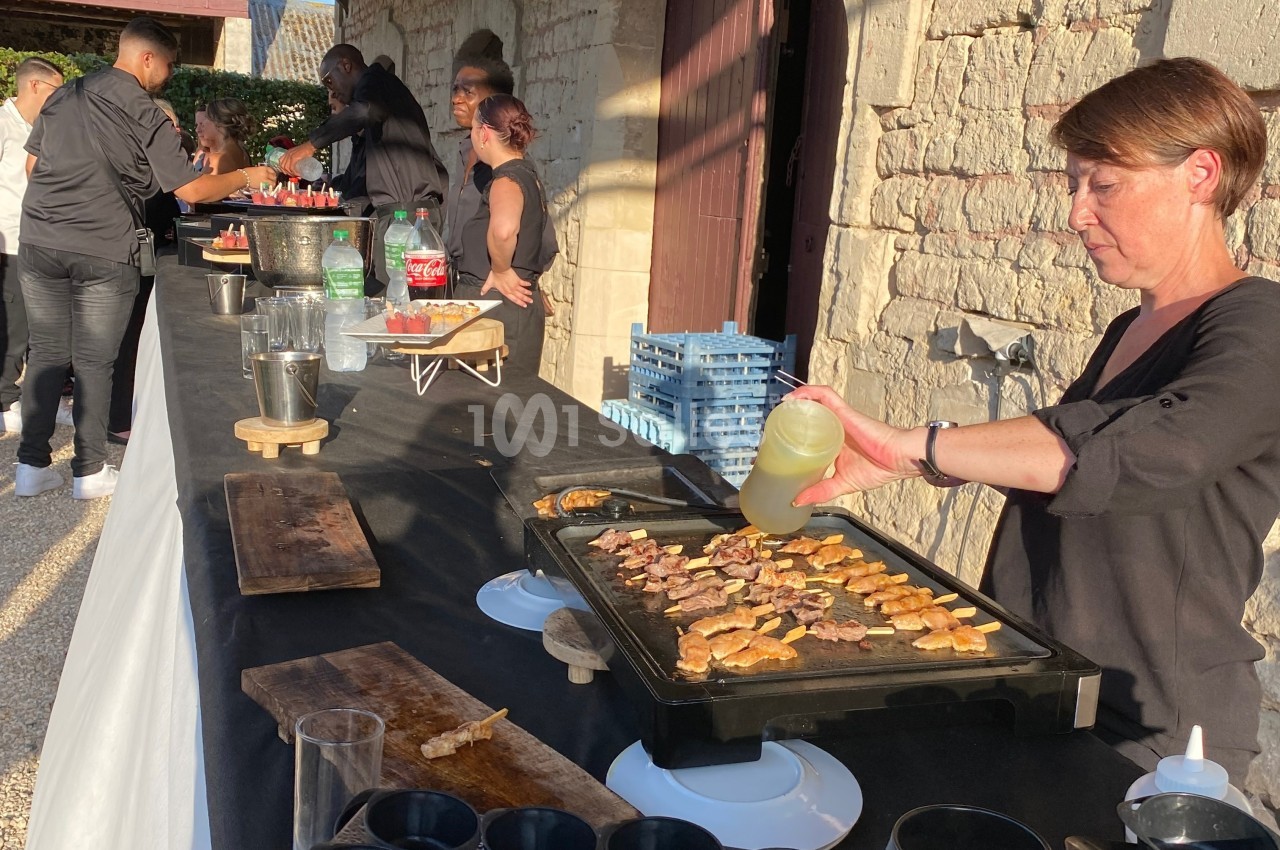 Une femme cuisine des aliments sur une plancha lors d'un buffet en extérieur, avec des serveurs en arrière-plan.