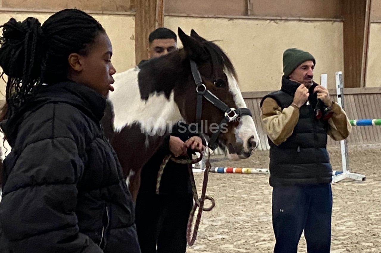 Deux personnes encadrent un cheval dans un manège équestre, une troisième ajuste son bonnet à l'arrière-plan.