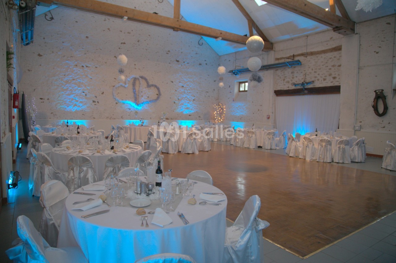 Salle de réception décorée avec des tables rondes, nappes blanches, éclairage bleu et piste de danse en bois.