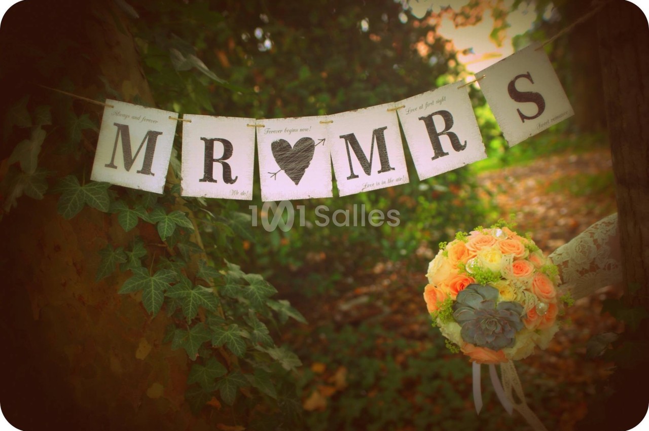Guirlande ’Mr & Mrs’ suspendue entre des arbres, avec un bouquet de roses pastel en premier plan.