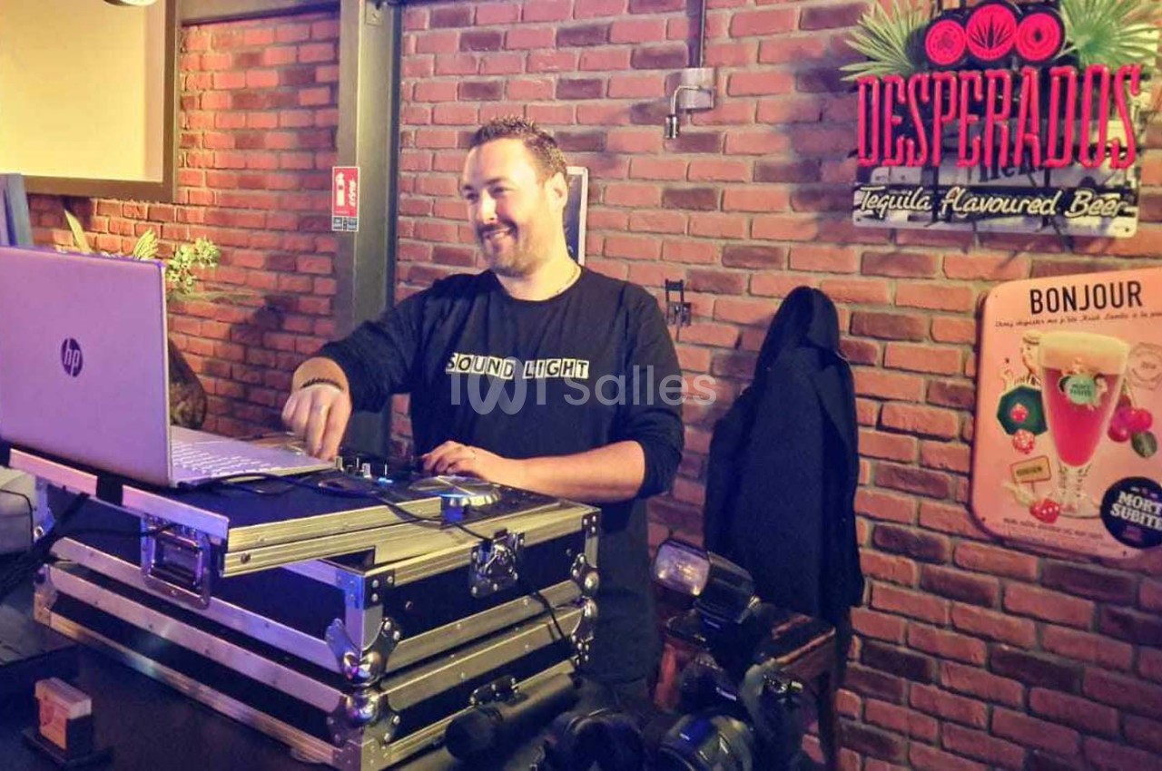 Un DJ souriant mixe de la musique sur un ordinateur portable dans un bar à la décoration murale en briques.