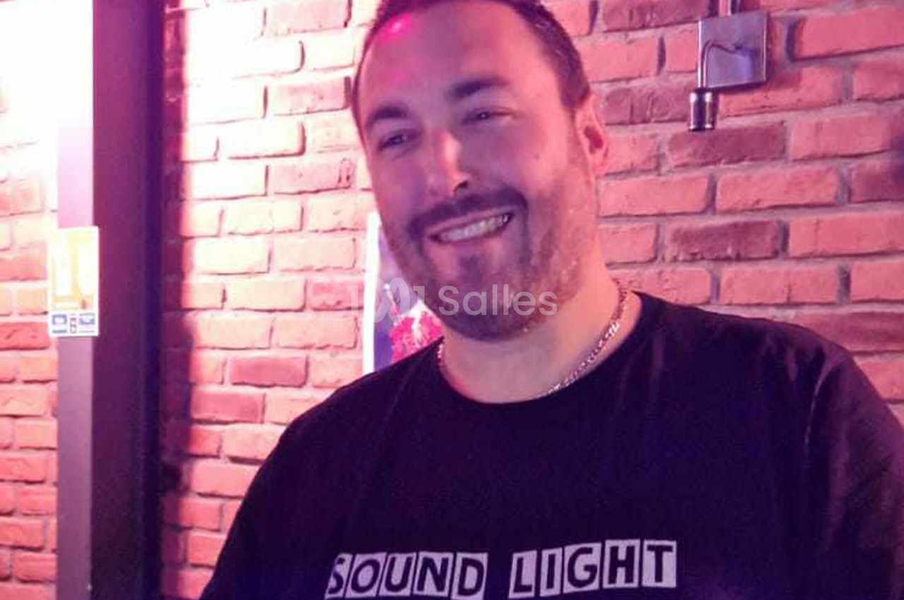 Homme souriant portant un t-shirt noir avec l'inscription ’Sound Light’, devant un mur de briques rouges.