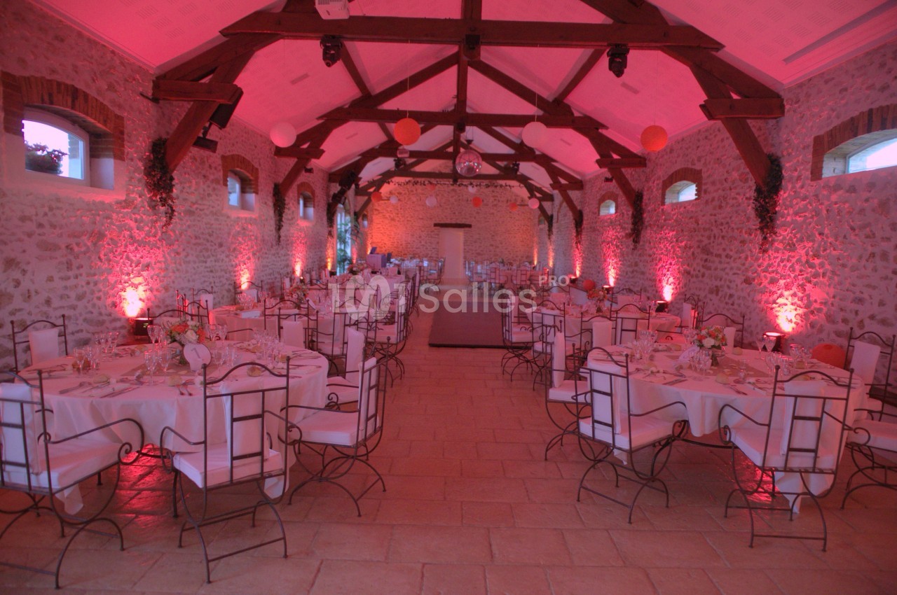 Salle de réception en pierre avec poutres apparentes, tables dressées et éclairage d'ambiance rose.
