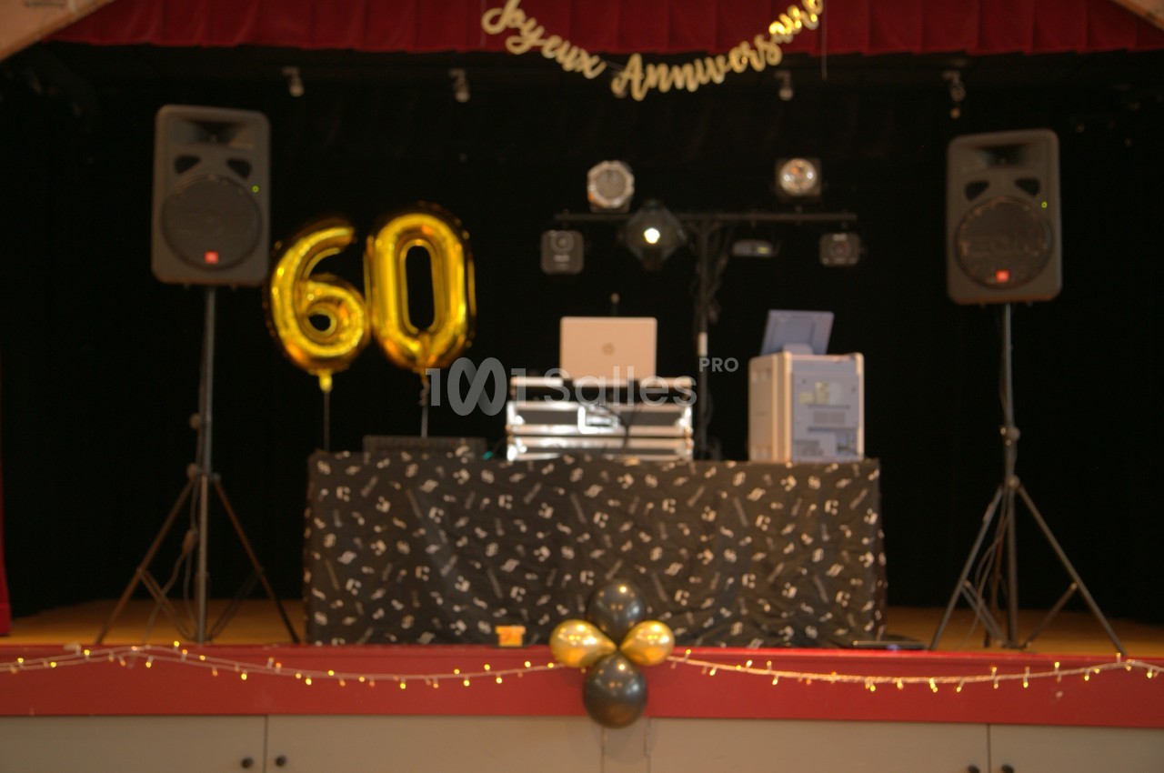 Table de DJ décorée pour un 60e anniversaire, avec ballons dorés, guirlandes lumineuses et équipements audio sur scène.