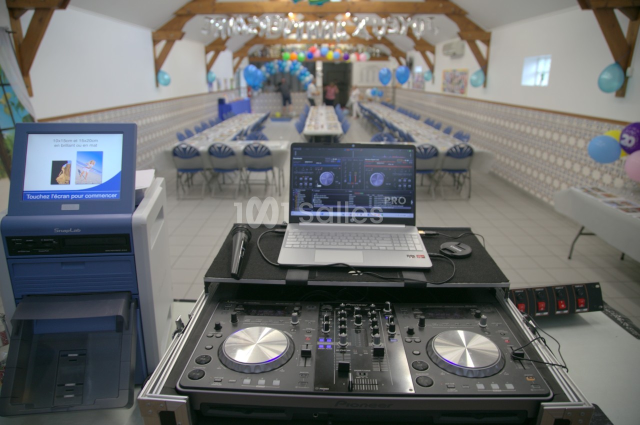 Table de mixage et ordinateur installés dans une salle décorée pour un événement avec tables alignées et ballons colorés.