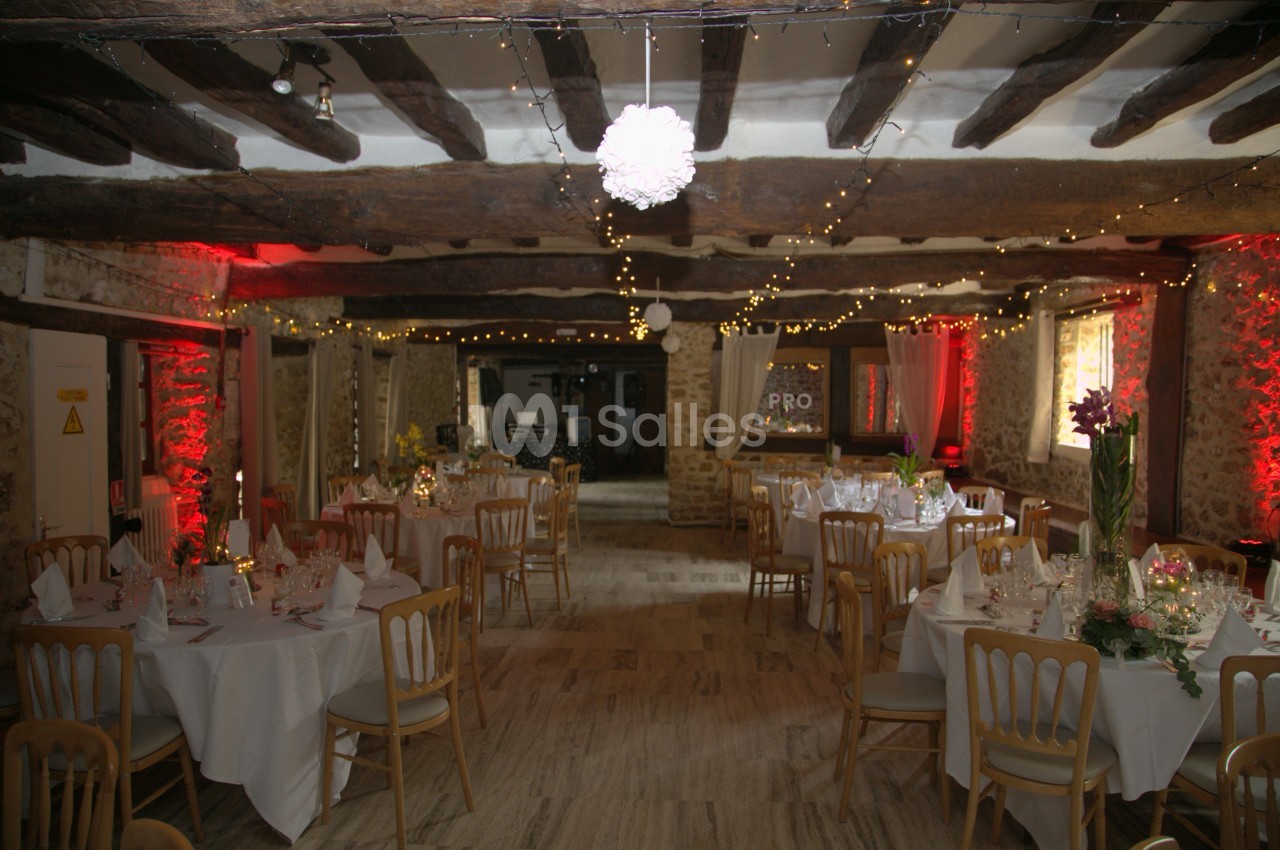 Salle de réception rustique décorée de guirlandes lumineuses, tables rondes dressées avec nappes blanches et chaises en bois.