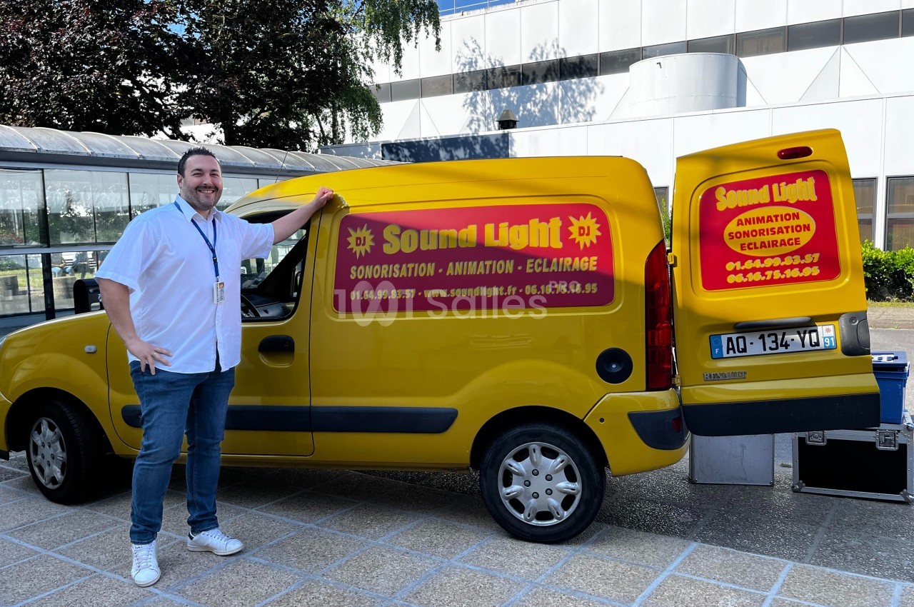 Un homme souriant se tient près d'un véhicule jaune marqué ’Sound Light’, avec du matériel audio visible à l'arrière.