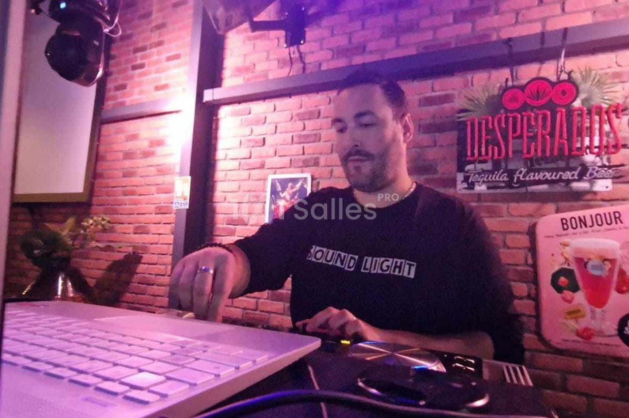 Un DJ mixe de la musique sur une console dans un bar à l'ambiance lumineuse et mur en briques.