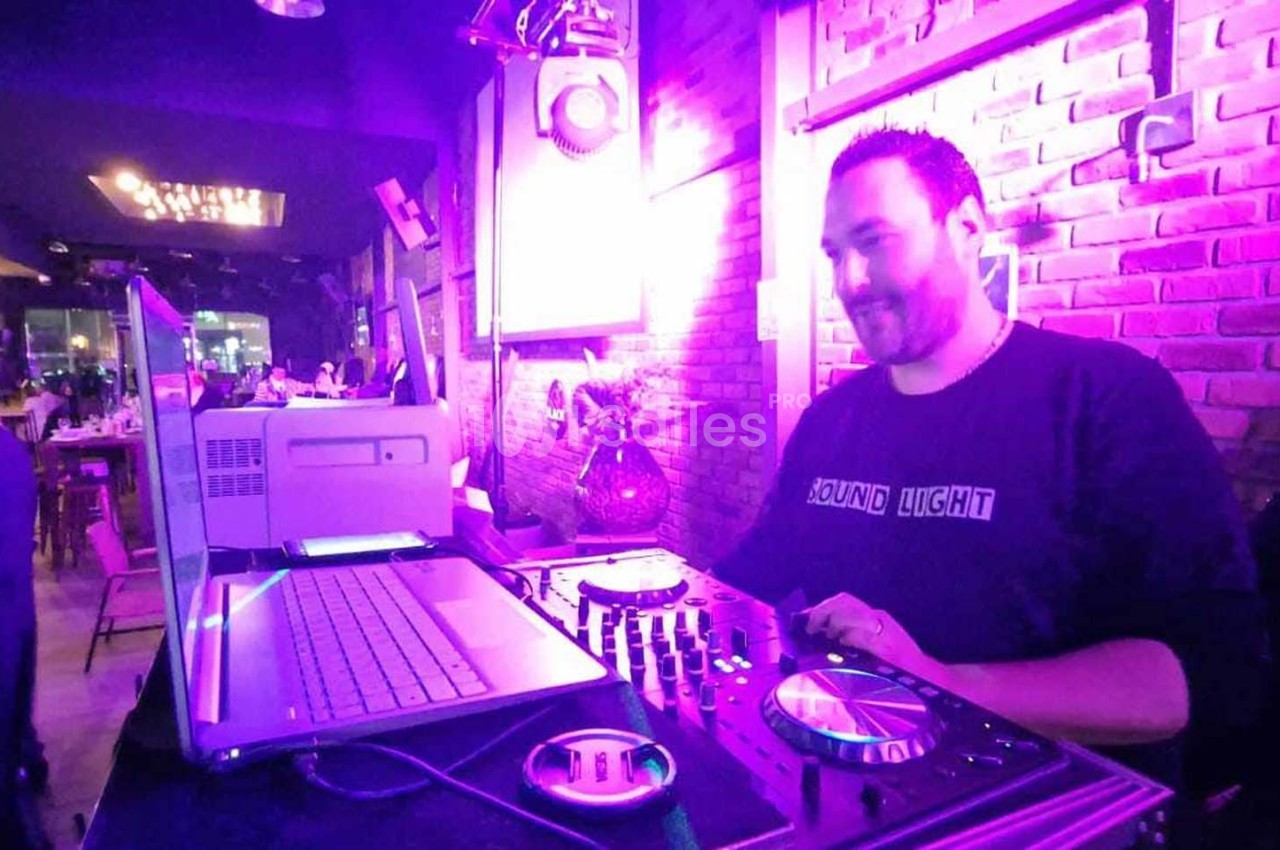 Un DJ mixe de la musique dans un bar éclairé par des lumières violettes, avec un ordinateur et une table de mixage.