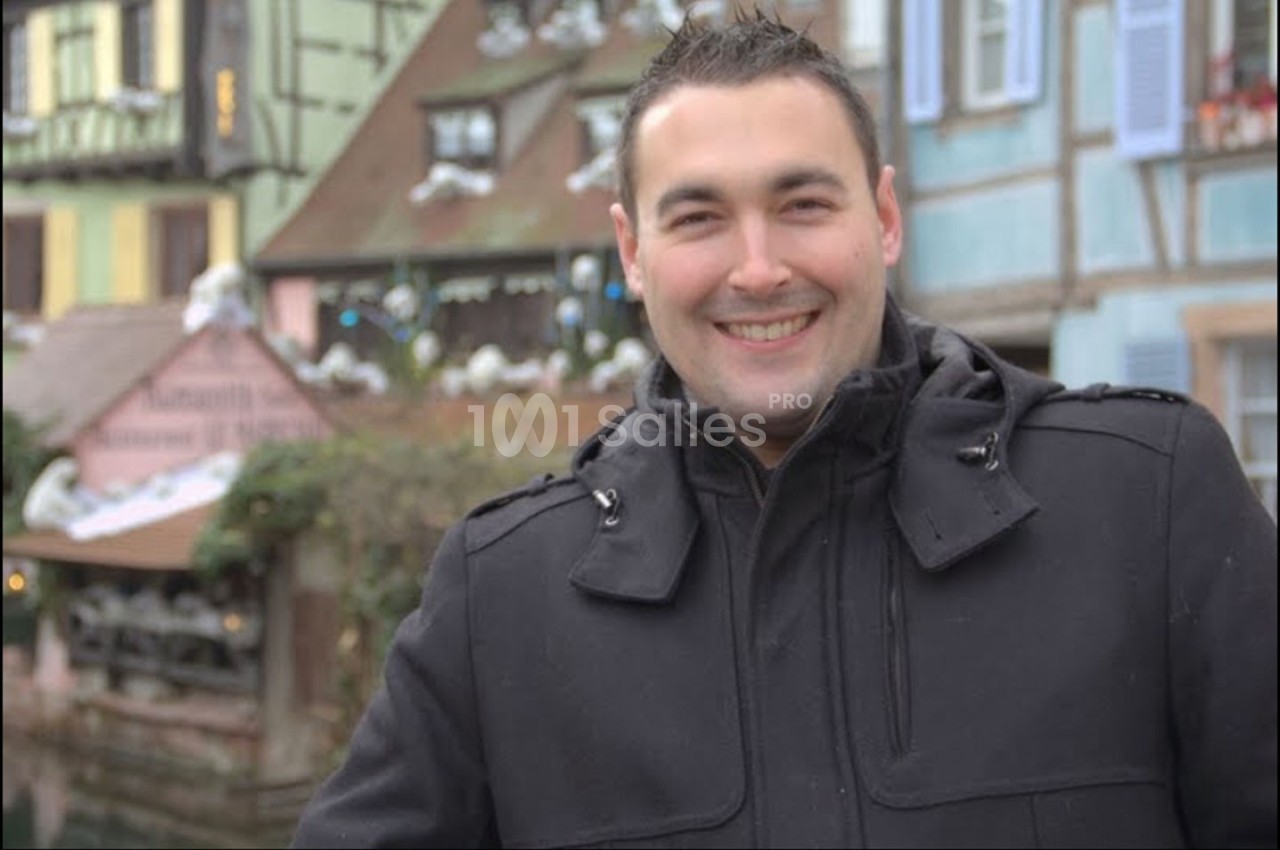Homme souriant en manteau noir devant des maisons à colombages colorées dans un décor urbain hivernal.
