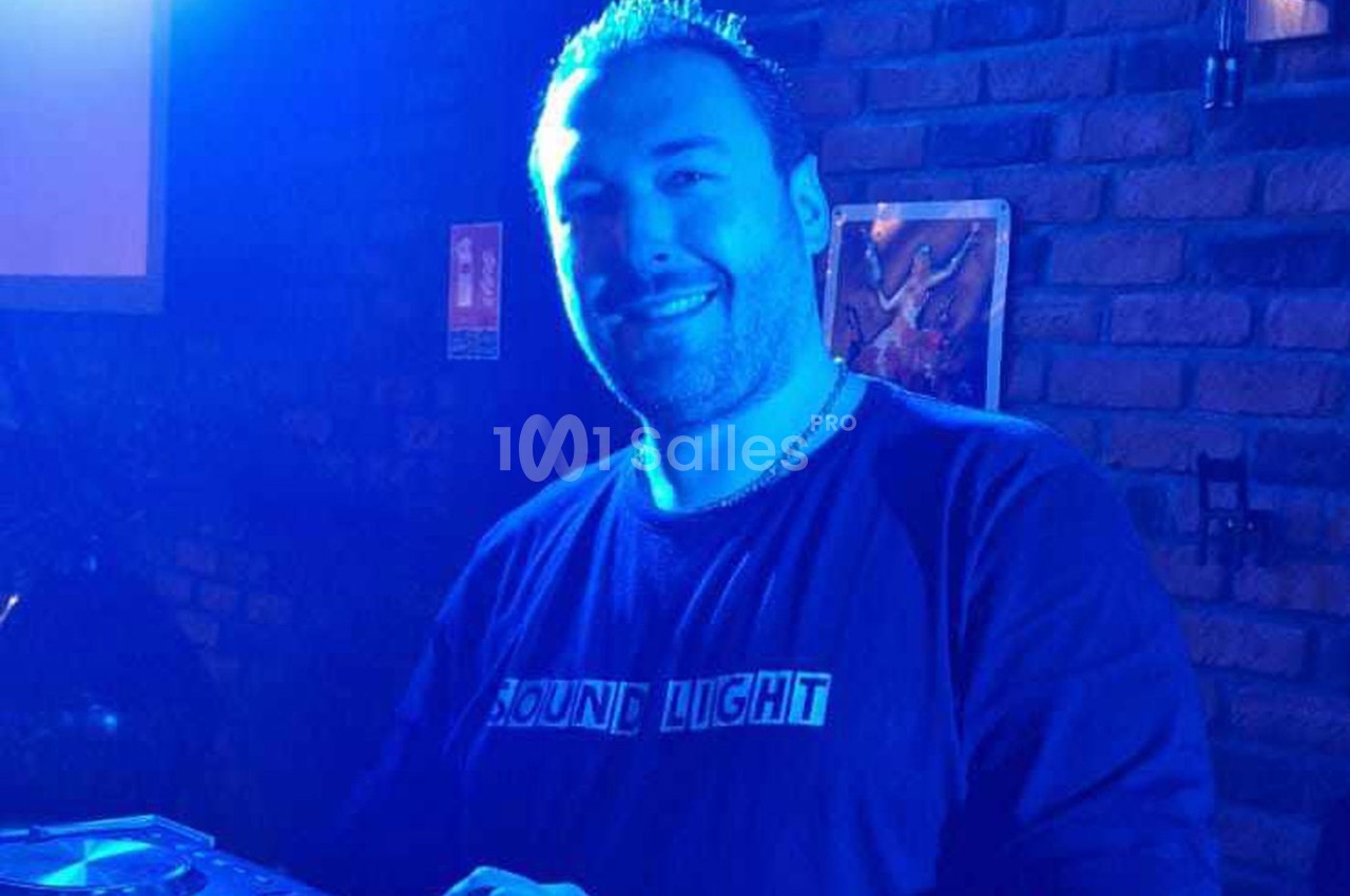 Homme souriant dans une ambiance lumineuse bleutée, debout devant un mur de briques avec un équipement audio visible.