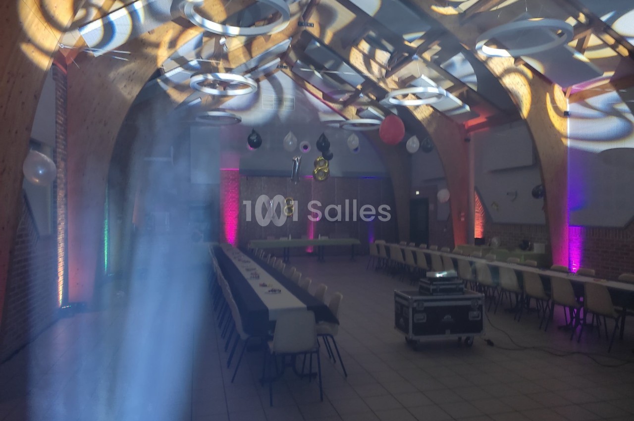 Salle décorée pour un événement, avec tables alignées, éclairages colorés et ballons suspendus au plafond.