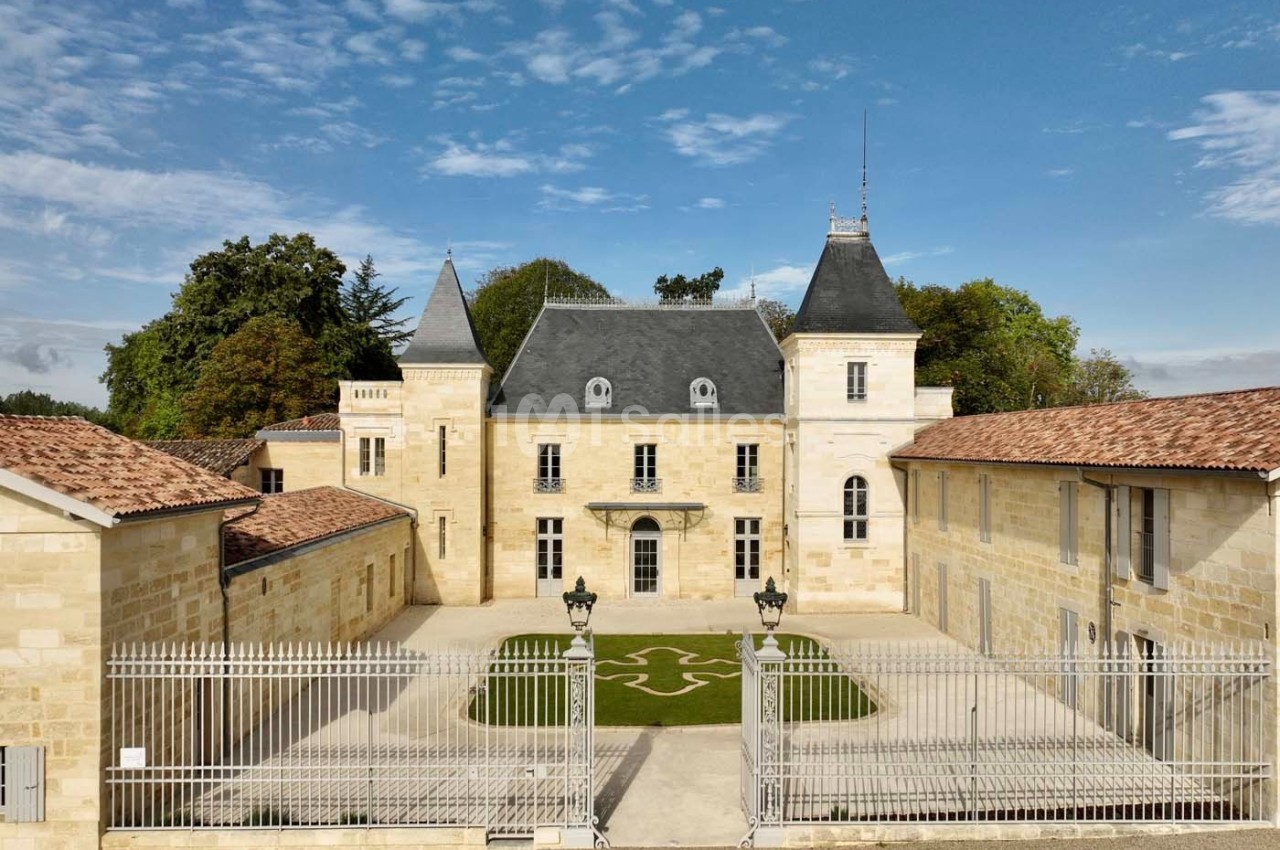 Façade d'un château en pierre entouré de bâtiments annexes, avec une grille en premier plan et un ciel dégagé.