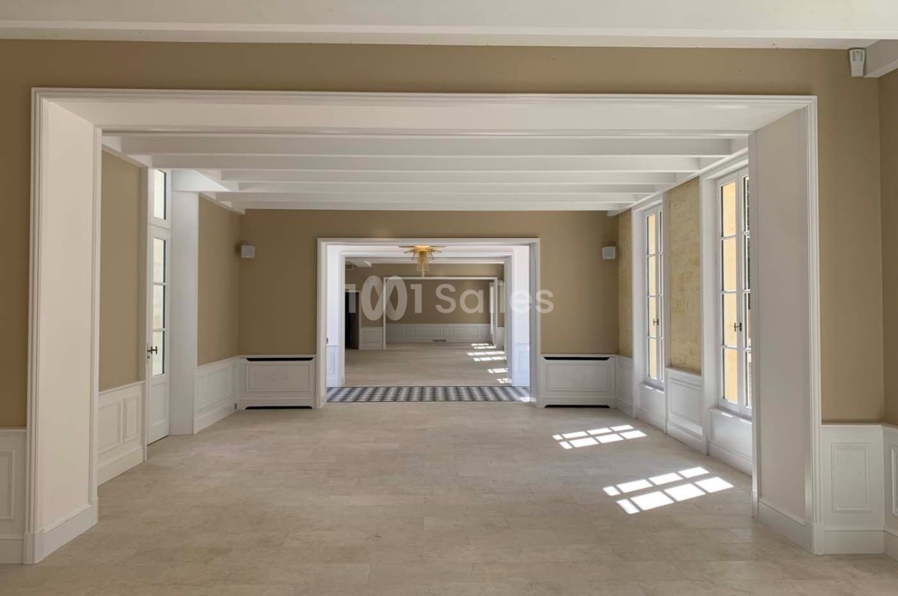 Salle lumineuse avec murs beiges, poutres blanches au plafond, grandes fenêtres et sol en pierre claire.