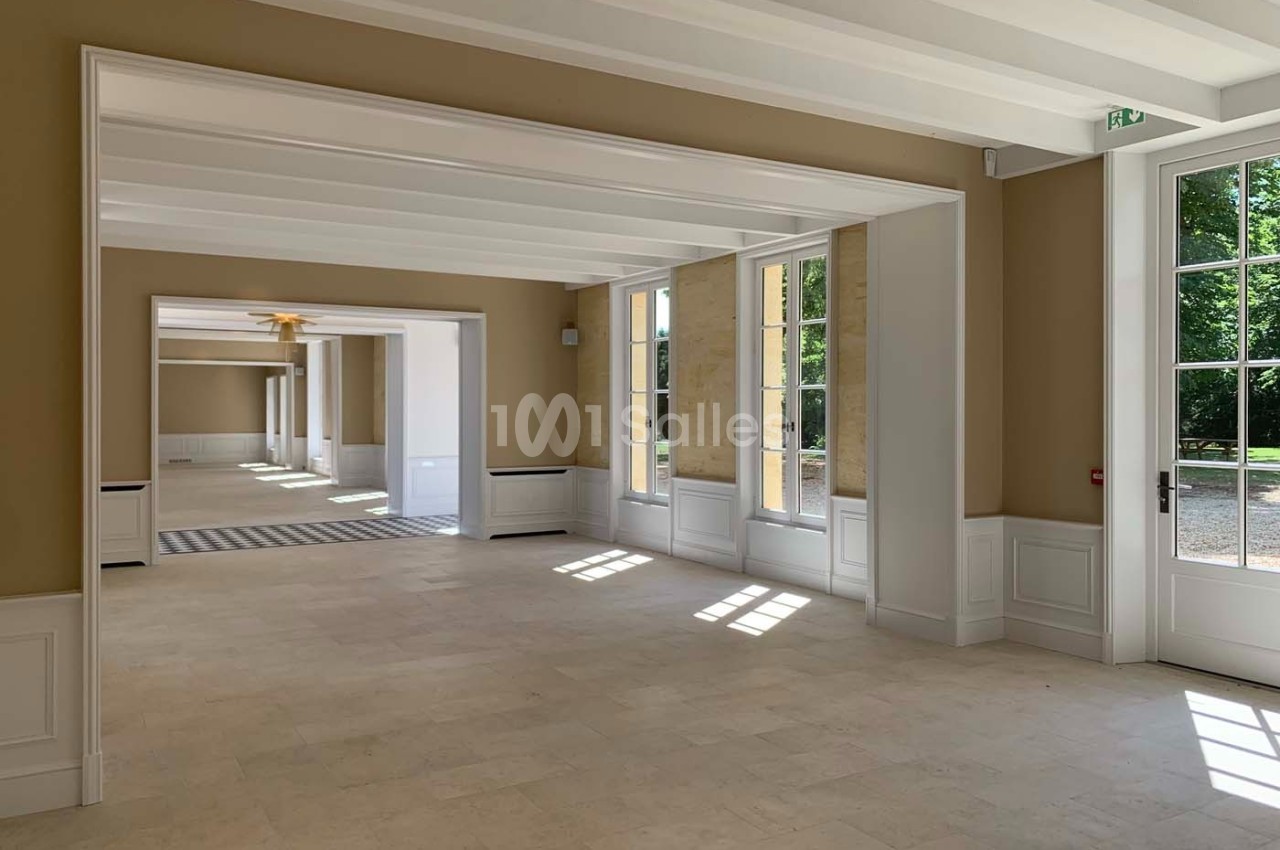 Intérieur lumineux avec grandes fenêtres, murs beige clair, sol en pierre et vue sur un jardin verdoyant.