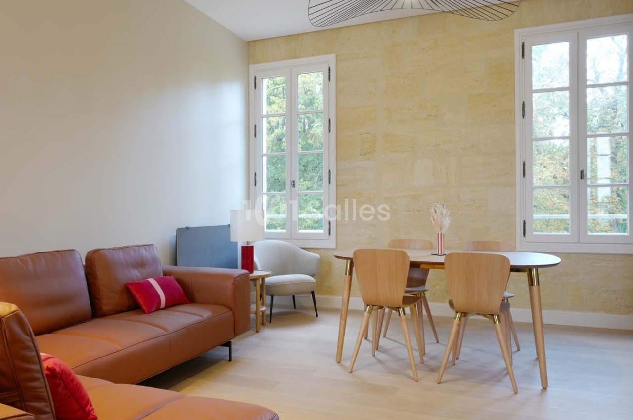 Salon lumineux avec murs en pierre, canapé en cuir, table en bois entourée de chaises et grandes fenêtres donnant sur l…