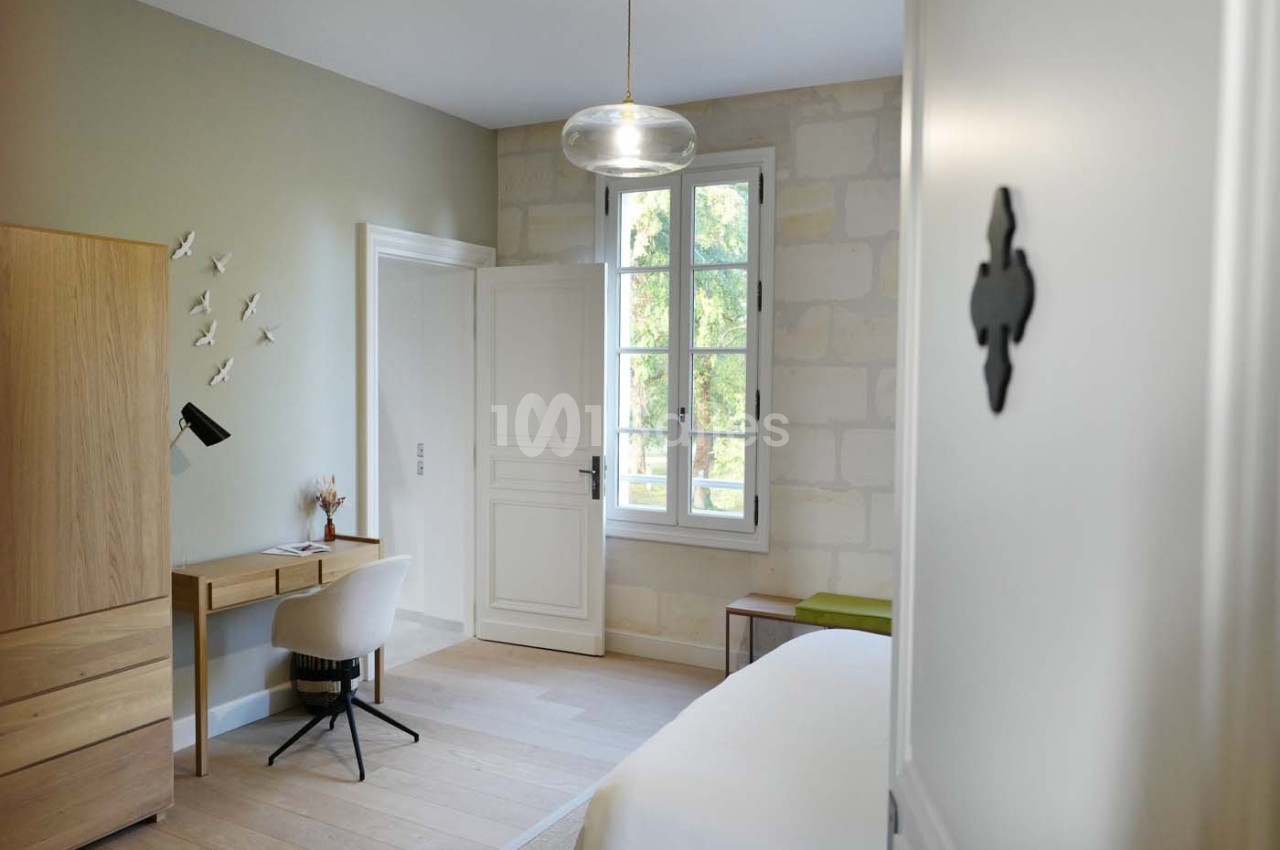 Chambre lumineuse avec bureau en bois, chaise blanche, armoire, lit et fenêtre donnant sur un extérieur verdoyant.