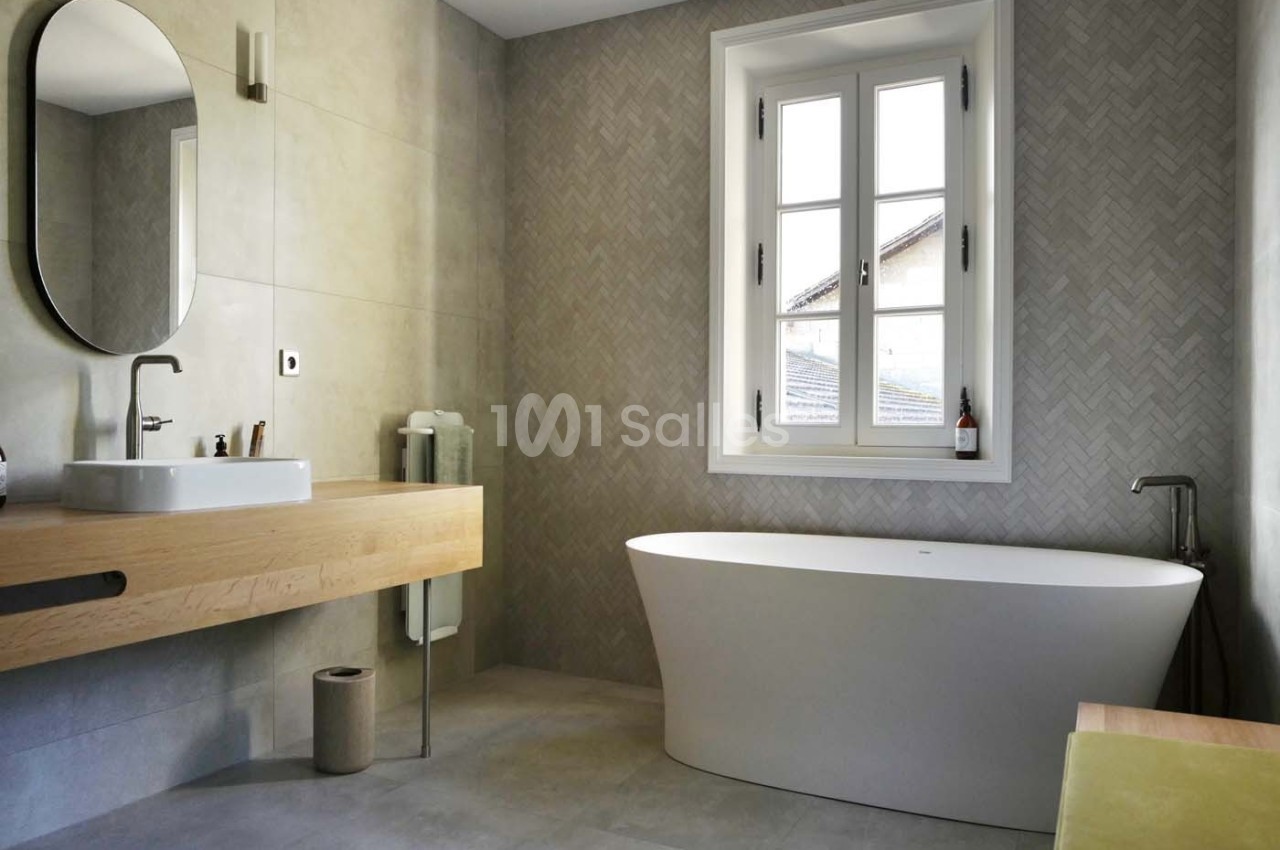 Salle de bain moderne avec baignoire îlot, vasque sur meuble en bois, miroir ovale et fenêtre à double battant.