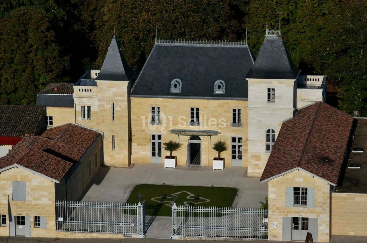 Façade d'un château en pierre avec deux tours, entouré de bâtiments annexes et d'une grille en fer forgé.