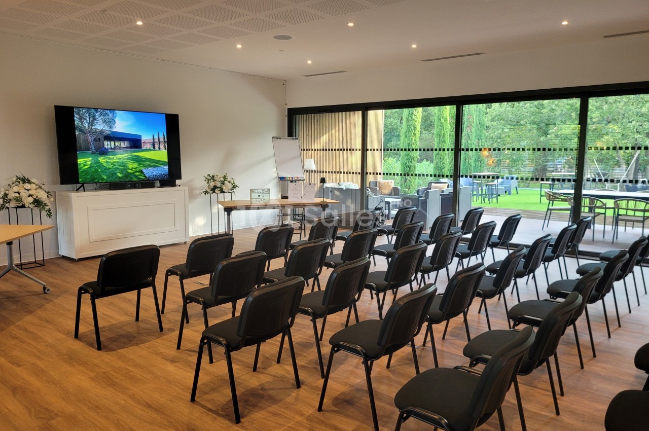 Salle de conférence lumineuse avec chaises alignées, écran allumé et vue sur un jardin à travers de grandes baies vitrées.