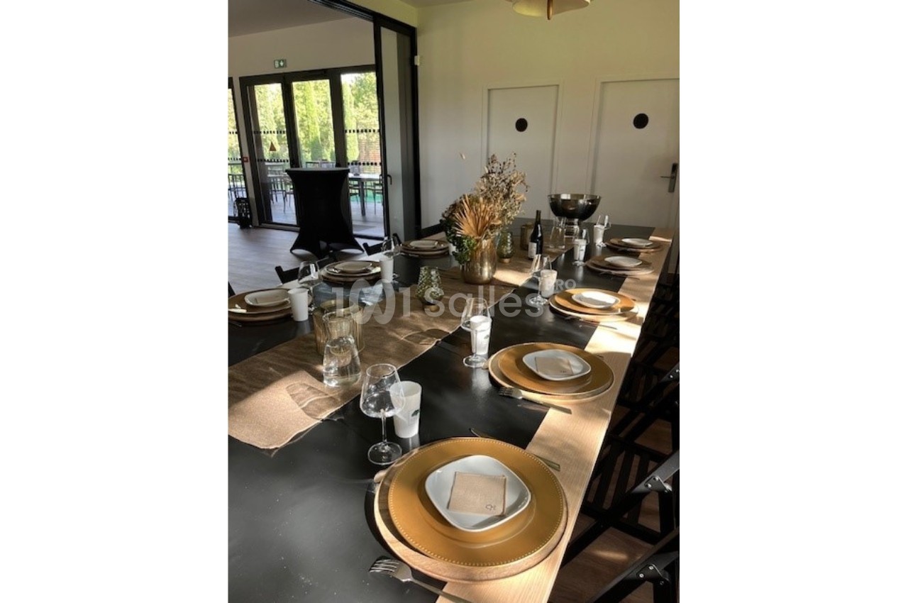 Table dressée avec vaisselle dorée, verres et décoration florale, dans une salle lumineuse avec vue sur l'extérieur.