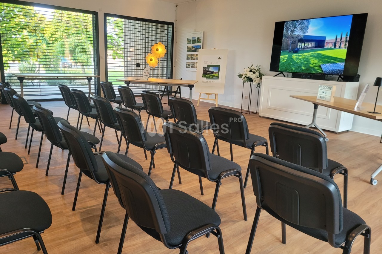 Salle de réunion lumineuse avec des rangées de chaises noires, un écran affichant une image et des présentoirs.