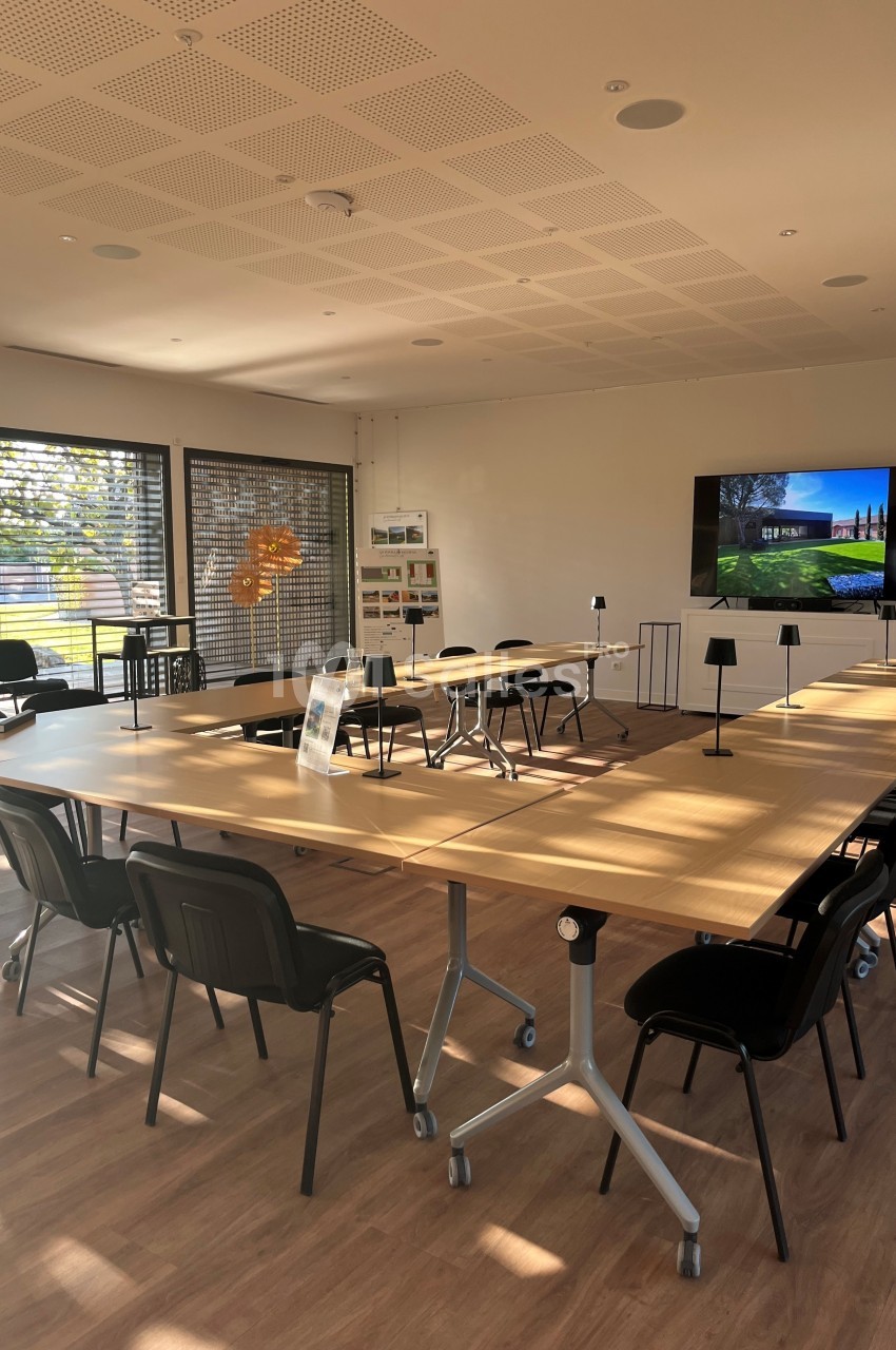 Salle de réunion lumineuse avec tables en bois disposées en U, chaises noires et écran affichant une image.